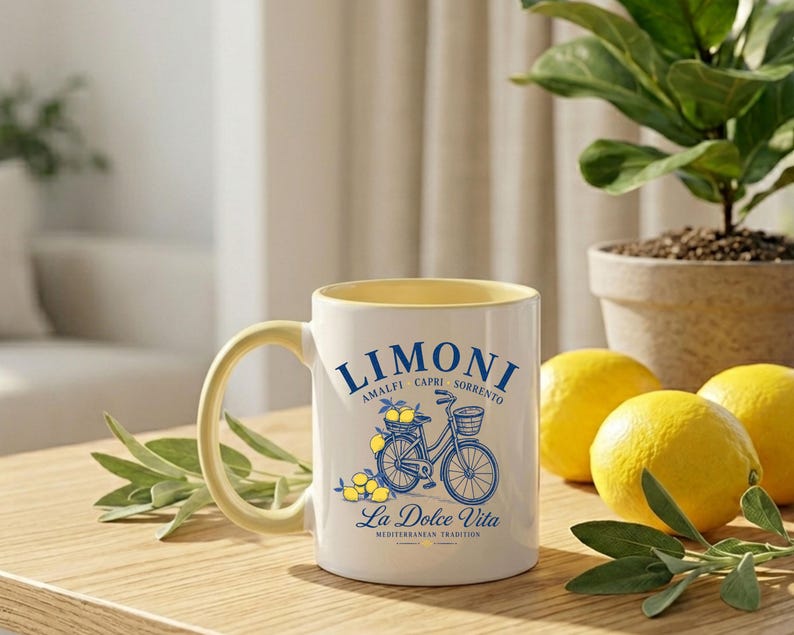 May include: A white ceramic mug with a pale yellow interior and handle. The front features a blue graphic of a bicycle with a basket of lemons, surrounded by the text LIMONI, AMALFI CAPRI SORRENTO, and La Dolce Vita MEDITERRANEAN TRADITION.