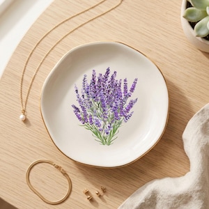 May include: A white ceramic trinket dish with a gold rim, featuring a lavender bouquet illustration. Gold jewelry, including a necklace, bracelet, and earrings, is arranged around the dish on a light wood surface. A small succulent in a white pot is also visible.