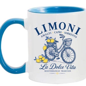 May include: A white ceramic mug with a blue interior and handle. The front features a blue graphic of a bicycle with a basket of yellow lemons, text reading LIMONI AMALFI CAPRI SORRENTO, La Dolce Vita, and MEDITERRANEAN TRADITION in blue ink.