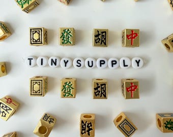 Puede incluir: Una colección de cuentas cuadradas de color dorado con varios caracteres chinos en negro, verde y rojo, dispuestas alrededor de una hilera central horizontal de cuentas redondas blancas que forman el texto TINY SUPPLY en letras negras.