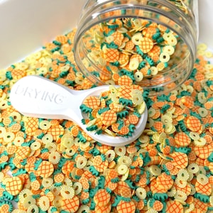 May include: A white scoop filled with yellow and orange pineapple and pineapple ring sprinkles. The sprinkles are in a clear jar with a white lid.