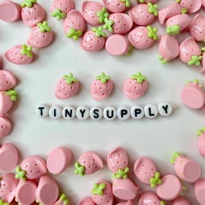 May include: A collection of small, light pink strawberry-shaped beads with green stems scattered around a single horizontal string of white round beads featuring the black printed text TINYSUPPLY in the center of the arrangement.