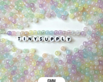 May include: A collection of small, round, translucent beads in pastel shades of pink, yellow, blue, and purple. A single strand of these beads is arranged horizontally above white letter beads spelling TINYSUPPLY. A label indicates 6MM.