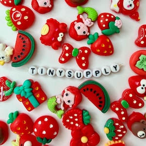 May include: A collection of colorful resin cabochons featuring strawberries, watermelons, mermaids, crabs, bows, and gift boxes, centered around a white string of letter beads spelling TINYSUPPLY in black font against a plain white background.