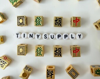 May include: A collection of gold-colored square beads featuring various Chinese characters in red, green, and black ink, arranged around a central horizontal string of white round beads spelling out the text TINY SUPPLY in black capital letters.