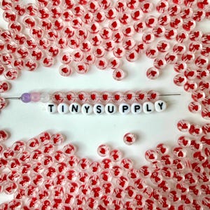 May include: A collection of clear round beads featuring small red heart patterns, scattered around a central needle threaded with a row of these heart beads, three purple beads, and a row of white letter beads spelling out TINYSUPPLY.