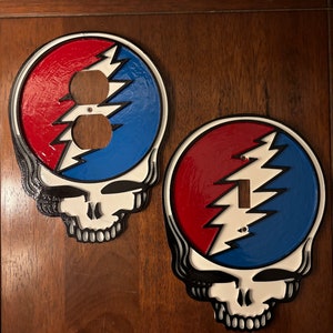 May include: Two decorative wall plates featuring a skull with a lightning bolt design. The plates are painted in red, white, and blue.