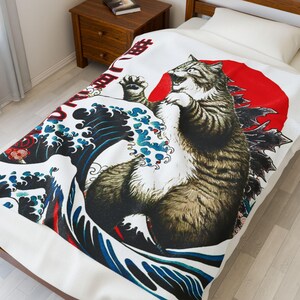 May include: White blanket featuring a tabby cat in a Godzilla-like pose against a red sun. The design incorporates Japanese wave patterns in blue and white, with Japanese text at the top. The blanket is on a bed.