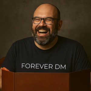 Può includere: Una persona sorridente indossa una maglietta nera con la scritta bianca "FOREVER DM". La persona indossa occhiali neri e ha una barba folta. Lo sfondo è di colore scuro e neutro.