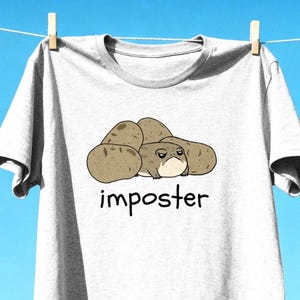 Puede incluir: Una camiseta gris claro de manga corta con un gráfico de una pequeña criatura redonda de color tostado con expresión gruñona escondida entre varias patatas marrones. La palabra "imposter" está impresa en letras negras, minúsculas y manuscritas.