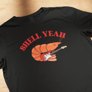 Könnte beinhalten: Ein schwarzes T-Shirt mit kurzen Ärmeln und einer Grafik einer orangefarbenen Garnele, die eine rote E-Gitarre spielt. Über der Garnele ist der Text SHELL YEAH in einer fetten roten Schrift gedruckt. Das Motiv ist mittig auf der Brust platziert.