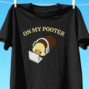 Könnte beinhalten: Ein schwarzes Kurzarm-T-Shirt mit einer Grafik eines braun-beigen Asseltiers, das graue Kopfhörer trägt und einen weißen Laptop benutzt. Über der Illustration ist der Text ON MY POOTER in einer hellgelben, serifenlosen Schrift gedruckt.