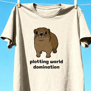 Cute brown hyrax illustration wearing a serious expression with the text “plotting world domination” printed on a t-shirt, funny animal meme graphic tee
