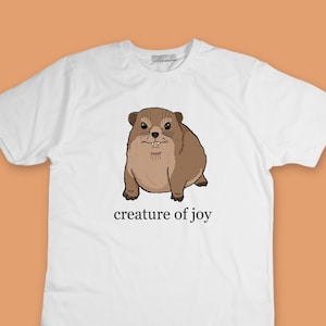 Puede incluir: Camiseta blanca con una ilustración de dibujos animados de un animal marrón y peludo y el texto "creature of joy". El animal tiene un cuerpo redondo, orejas pequeñas y dientes visibles. El texto es en negro, sin serifa. La camiseta se muestra sobre un fondo naranja claro.