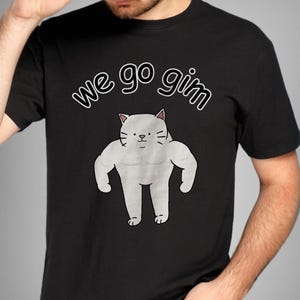 Könnte beinhalten: Ein schwarzes T-Shirt mit kurzen Ärmeln und einer Grafik einer weißen, muskulösen Katze mit übertrieben großen Armen. Über der Katze steht der Text "we go gim" in weißer Schrift mit schwarzer Umrandung. Das Motiv ist mittig platziert.