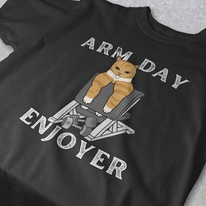 May include: Black t-shirt featuring a cartoon cat with orange and white stripes, lifting weights on a bench. The text "ARM DAY ENJOYER" is printed in white above and below the cat illustration.