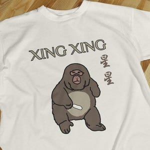 May include: A white t-shirt featuring a graphic of a brown gorilla with a light tan belly, holding a small white object. The text "XING XING" is printed in large dark letters above the gorilla, with two Chinese characters to the right of the figure.