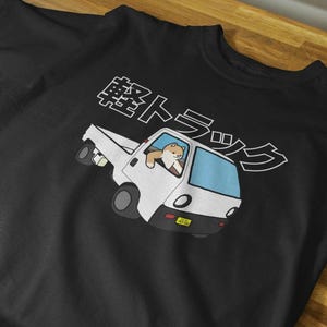 May include: A black t-shirt featuring a graphic of a white kei truck with a light blue windscreen. A brown and white dog is visible in the driver's seat. Above the truck, the Japanese text 軽トラック is printed in white with a black outline.