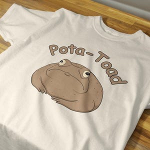 Puede incluir: Camiseta blanca con una ilustración de dibujos animados de un sapo marrón y el texto "Pota-Toad" en marrón. El sapo tiene ojos grandes y una expresión gruñona. La camiseta está hecha de una tela suave y clara.