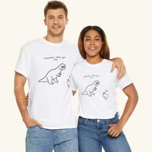 May include: A white short-sleeved t-shirt featuring a black line drawing of a Tyrannosaurus Rex and a small chicken. The text "remember who you are" is printed above the dinosaur, and the word "bok" is printed near the chicken.