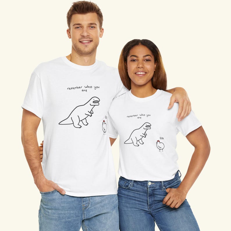 May include: A white short-sleeved t-shirt featuring a black line drawing of a Tyrannosaurus Rex and a small chicken. The text "remember who you are" is printed above the dinosaur, and the word "bok" is printed near the chicken.