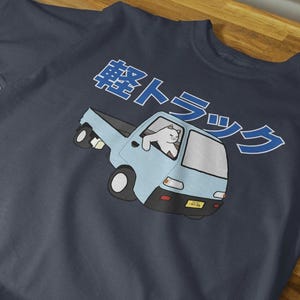 Puede incluir: Una camiseta azul marino con una ilustración de dibujos animados de un gato blanco conduciendo una camioneta azul. El texto "軽トラック" está impreso en azul en la camiseta.