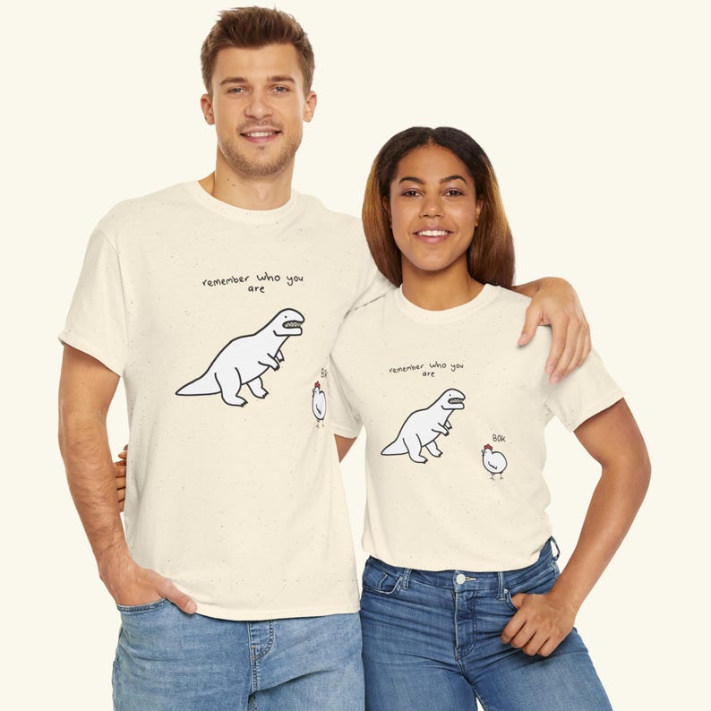 May include: A cream-colored short-sleeve t-shirt with a speckled texture. The graphic features a line-art illustration of a dinosaur looking at a chicken, with the text "remember who you are" above the dinosaur and "bok" near the chicken.
