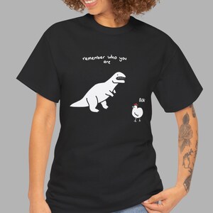 May include: A black short-sleeve t-shirt featuring a white graphic of a dinosaur and a chicken. Above the dinosaur, the text reads "remember who you are" in white. Next to the chicken, the text reads "BOK" in white with a small red comb.