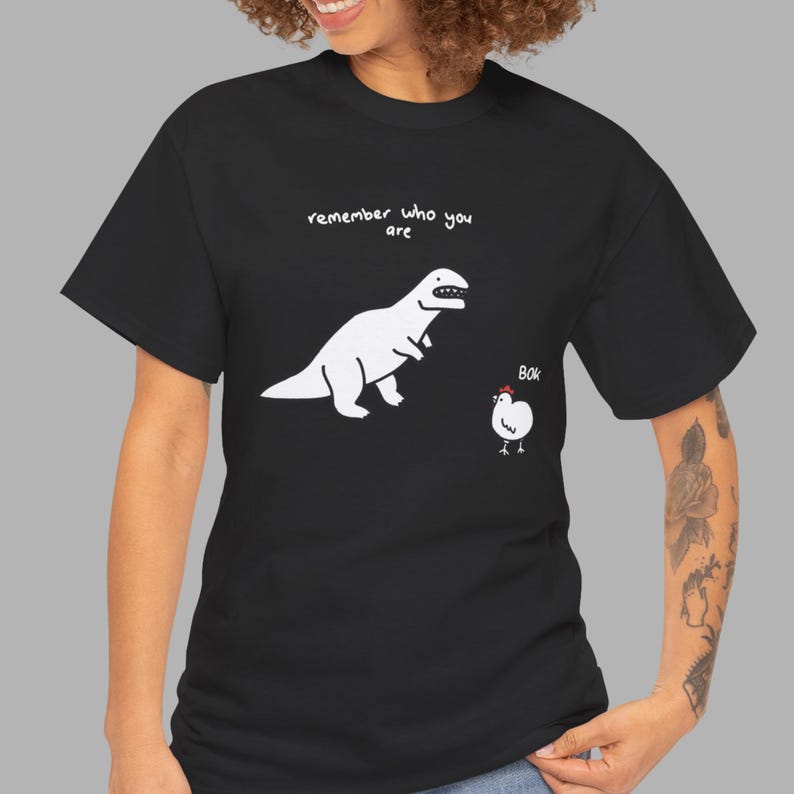 May include: A black short-sleeve t-shirt featuring a white graphic of a dinosaur and a chicken. Above the dinosaur, the text reads "remember who you are" in white. Next to the chicken, the text reads "BOK" in white with a small red comb.