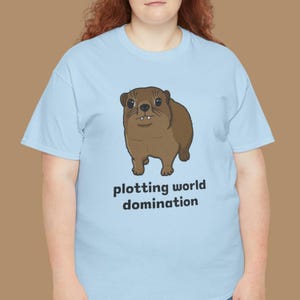 Cute brown hyrax illustration wearing a serious expression with the text “plotting world domination” printed on a t-shirt, funny animal meme graphic tee