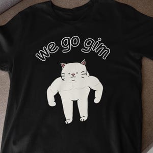 Könnte beinhalten: Ein schwarzes kurzärmeliges T-Shirt mit einer Grafik einer weißen, muskulösen Katze mit kleinem Gesicht und dem Text "we go gim" in weißen Buchstaben über der Katze. Das Design ist mittig auf der Brust des Kleidungsstücks platziert.