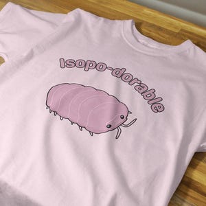 Puede incluir: Camiseta rosa claro con un dibujo de un isópodo rosa y el texto "isopo-dorable" encima. El isópodo tiene un cuerpo segmentado, ojos pequeños y antenas. La camiseta es de algodón.