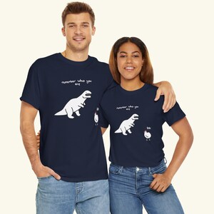 May include: Two navy blue short-sleeve t-shirts featuring a white graphic of a T-Rex looking at a small chicken. The text "remember who you are" is printed above the dinosaur, and the word "Bok" is printed next to the chicken on both shirts.