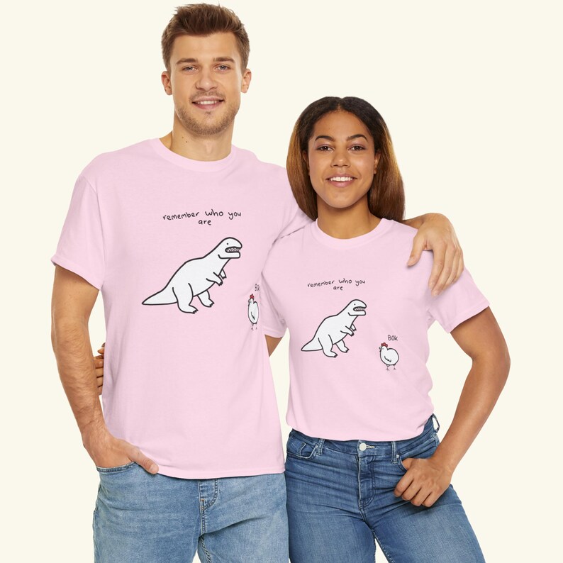 May include: A light pink t-shirt featuring a black line drawing of a dinosaur and a chicken. Above the dinosaur is the text "remember who you are" and next to the chicken is the text "bok". The design is printed on the front of the garment.