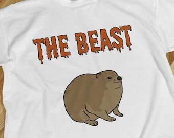 Könnte beinhalten: Weißes T-Shirt mit dem Text "THE BEAST" in orangefarbenen, tropfenden Buchstaben. Darunter eine Cartoon-Illustration eines braunen, stämmigen Tieres mit ernstem Gesichtsausdruck.