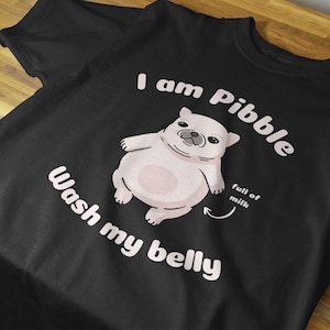 May include: Black t-shirt featuring a cartoon illustration of a light-coloured, chubby animal. The text reads "I am Pibble" and "Wash my belly." The phrase "full of milk" is also included.