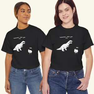 May include: A black short-sleeved t-shirt featuring a white graphic of a dinosaur facing a chicken. Above the dinosaur is the text "remember who you are" and next to the chicken is the text "Bok". The shirt has a crew neckline and a straight hem.
