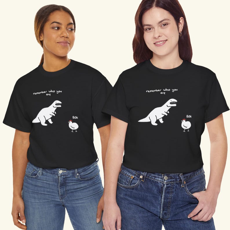 May include: A black short-sleeved t-shirt featuring a white graphic of a dinosaur facing a chicken. Above the dinosaur is the text "remember who you are" and next to the chicken is the text "Bok". The shirt has a crew neckline and a straight hem.