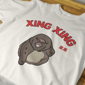 May include: A white t-shirt featuring a graphic of a brown, round-bodied primate with a light-colored belly and a wide, toothy grin. Above the illustration, the text XING XING is printed in red, with the Chinese characters 星星 printed below it.
