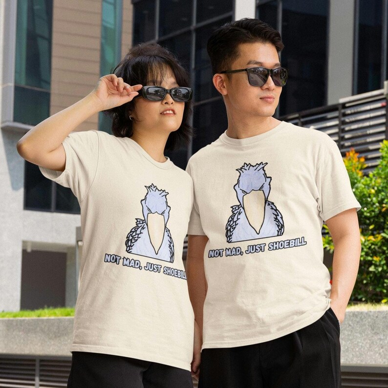 May include: A cream-colored short-sleeve t-shirt featuring a graphic illustration of a shoebill bird with a light blue body and a large beige beak. Below the bird, the text reads: NOT MAD, JUST SHOEBILL in a bold, dark blue font.