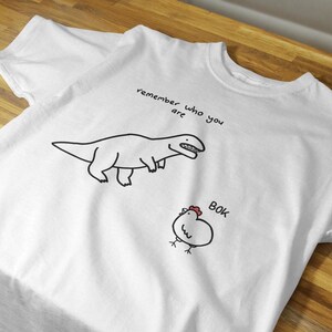 May include: A white short-sleeved t-shirt featuring a black line drawing of a dinosaur and a chicken. Above the dinosaur, the text reads "remember who you are" and next to the chicken, the text reads "BOK" in black with a small red comb.