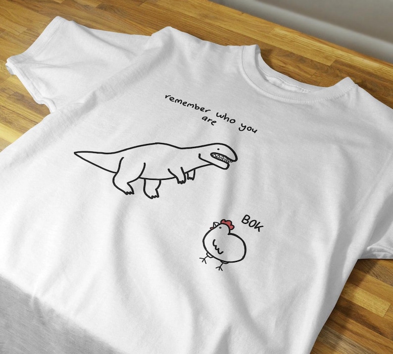 May include: A white short-sleeved t-shirt featuring a black line drawing of a dinosaur and a chicken. Above the dinosaur, the text reads "remember who you are" and next to the chicken, the text reads "BOK" in black with a small red comb.