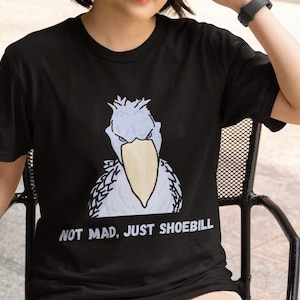 May include: A black short-sleeve t-shirt featuring a graphic illustration of a light blue shoebill stork with a large yellow beak. Below the bird, the text reads in white capital letters: NOT MAD, JUST SHOEBILL. The shirt has a crew neckline.