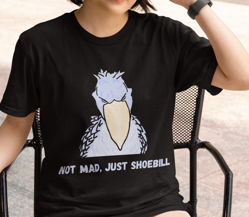 May include: A black short-sleeve t-shirt featuring a graphic illustration of a light blue shoebill stork with a large yellow beak. Below the bird, the text reads in white capital letters: NOT MAD, JUST SHOEBILL. The shirt has a crew neckline.