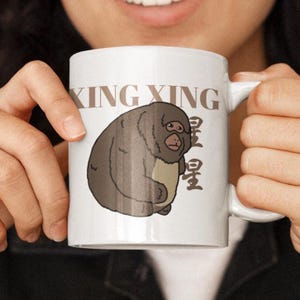 May include: White ceramic mug featuring a cartoon illustration of a monkey with the text "XING XING" in a brown font. The monkey is brown and beige. The mug has a handle on the right side.