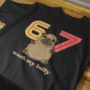 Pode incluir: Camiseta preta com um desenho de pug de desenho animado e o texto "wash my belly". O design inclui um número seis laranja e um símbolo de maior que rosa. O pug é marrom claro com detalhes em marrom escuro.