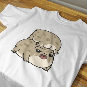 May include: White t-shirt featuring a cartoon illustration of two grumpy-looking frogs. The larger frog is on the bottom, with a smaller frog on top. The frogs are tan with dark spots and black outlines.