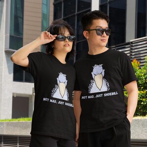 May include: A black short-sleeved t-shirt featuring a graphic illustration of a grey shoebill bird with a large beak. Below the bird, the text reads "NOT MAD, JUST SHOEBILL" in white capital letters. The shirt has a crew neckline and a straight hem.