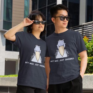 May include: A dark gray short-sleeved t-shirt featuring a graphic illustration of a shoebill bird with the text "NOT MAD, JUST SHOEBILL" printed in white capital letters below the bird. The design is centered on the front of the garment.