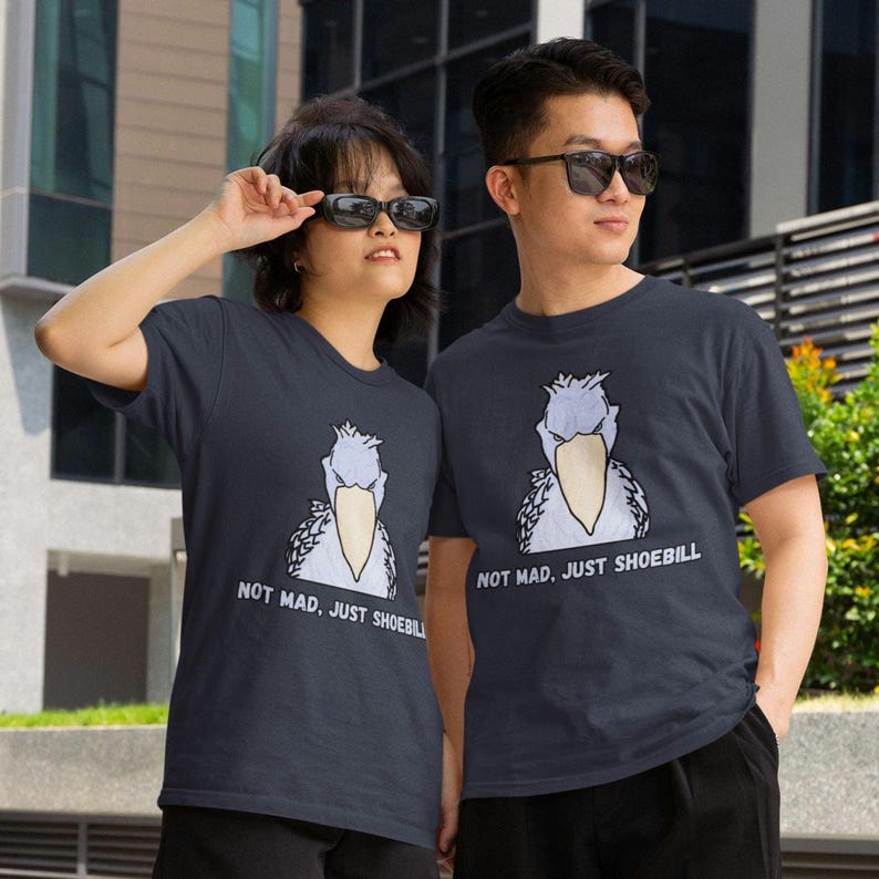 May include: A dark gray short-sleeved t-shirt featuring a graphic illustration of a shoebill bird with the text "NOT MAD, JUST SHOEBILL" printed in white capital letters below the bird. The design is centered on the front of the garment.
